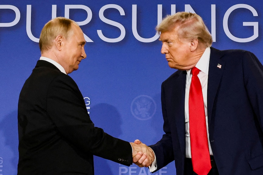 Two men in black suits shake hands in front of a blue backdrop