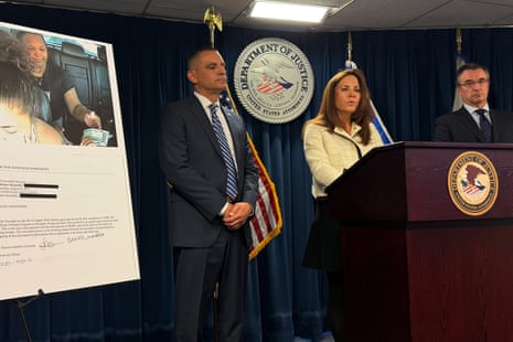 A woman speaks at a lectern between two men in front of blue curtain and seal marked 'Department of Justice'