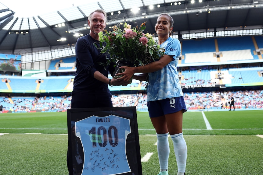 Australian footballer Mary Fowler smiles holding flowers in front of a shirt saying "Fowler 100".